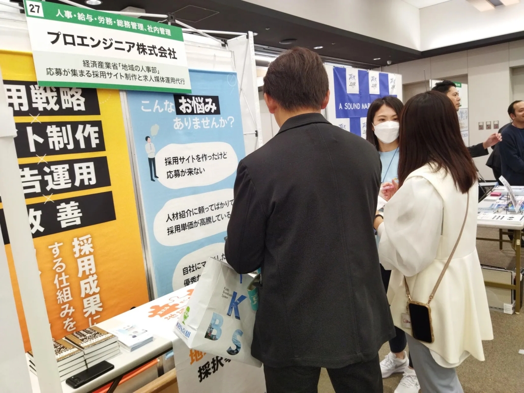 採用イベントのブースで、参加者が「プロエンジニア株式会社」の採用支援サービスについて説明を受けている様子。採用サイト制作や求人媒体運用代行に関する課題解決を提案しており、来場者とスタッフが交流している。