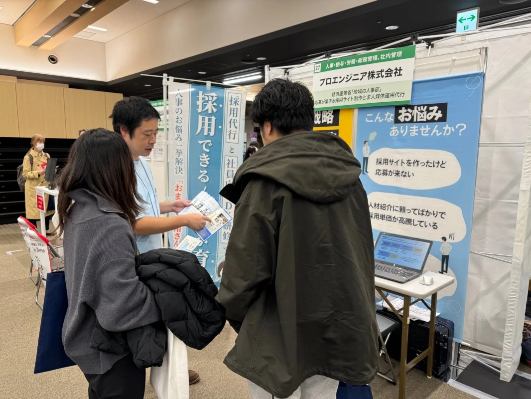 採用イベントのブースで、企業の担当者が来場者2名に資料を見せながら説明している様子。ブースには「採用できる」「こんなお悩みありませんか？」といった採用に関する課題解決を促すメッセージが掲示されている。