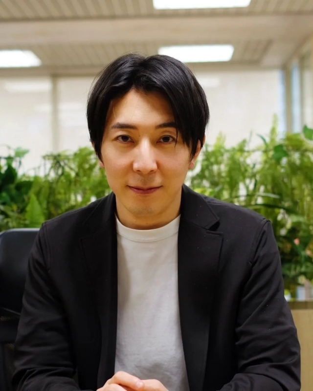 黒いジャケットを着た男性がカメラ目線で座っている写真です。背景には緑の植物と明るい照明が見えます。