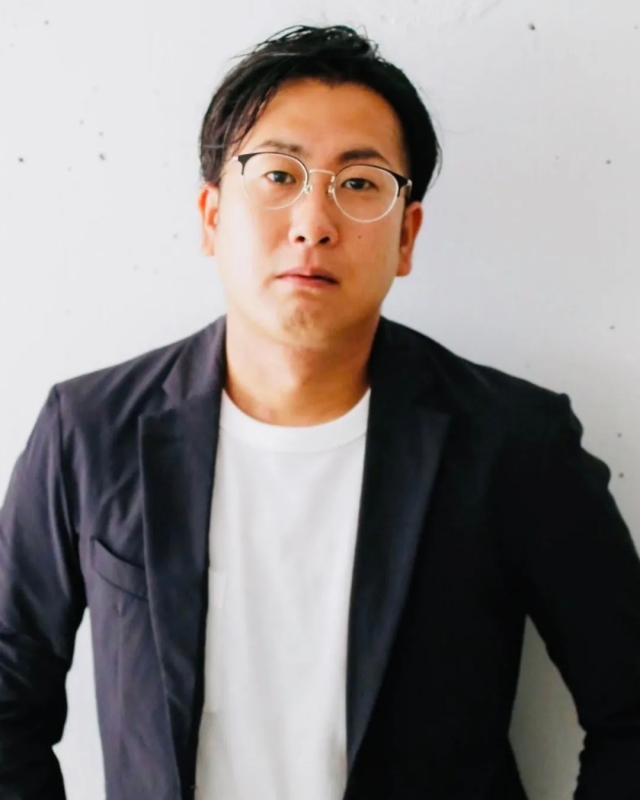 眼鏡をかけた男性がこちらを見ているポートレート写真です。彼は白いTシャツと黒いジャケットを着ています。