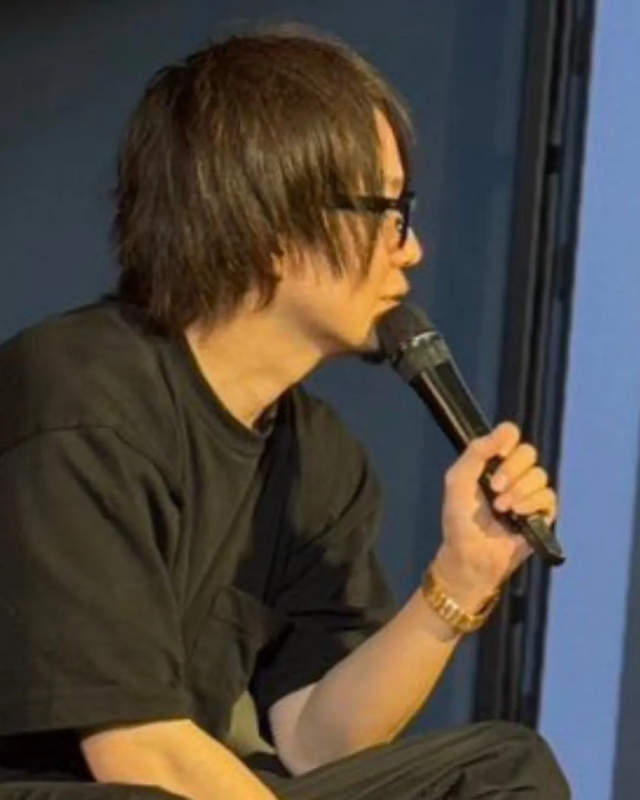 眼鏡をかけた男性がマイクを持って話している様子を捉えた写真です。彼は黒い服を着ており、腕には金色のブレスレットをしています。